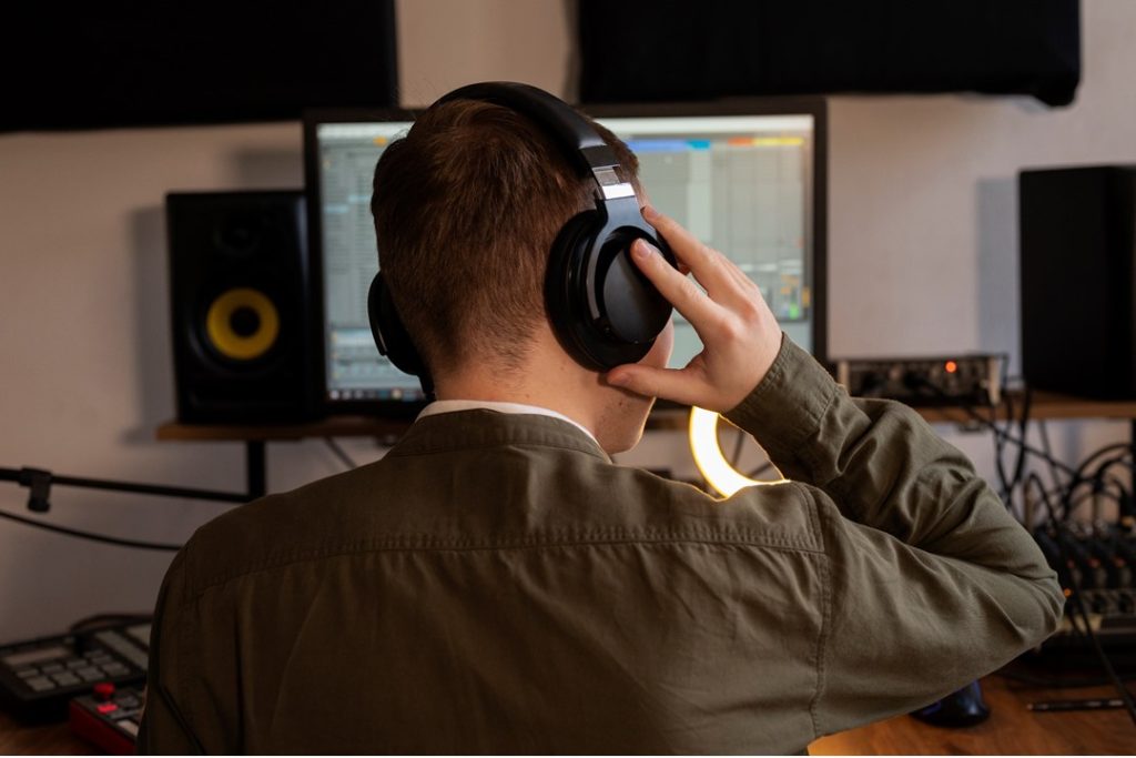 一位男生背對著鏡頭，面對電腦使用音樂剪輯軟體，並戴著耳機仔細聆聽音樂檔案