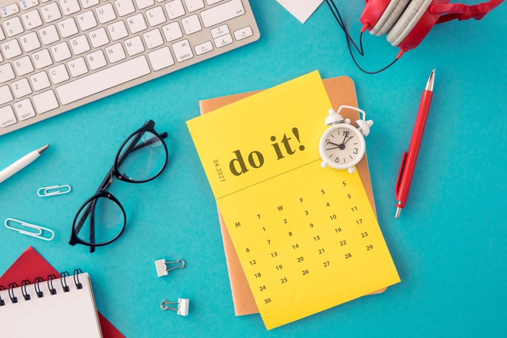 一張藍色的桌上，放著許多文具及辦公用品，中間黃色行事曆上寫著do it！的英文，執行就對了