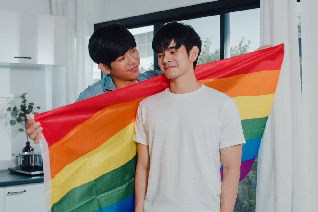 兩個男生在家裡，其中一人拿著象徵同志的彩虹旗，幫另一人披上，每個人都生而獨特