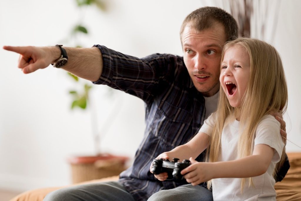 女孩拿著電動控制器開心的玩著，一旁女孩的爸爸也正認真的指導著